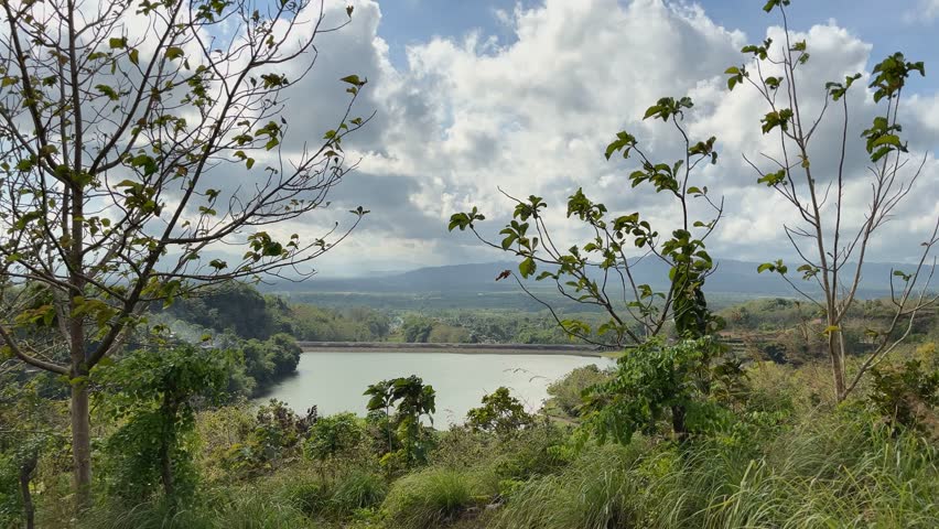 Aerial view of lake surrounded by green hills and forest under bright cloudy sky.