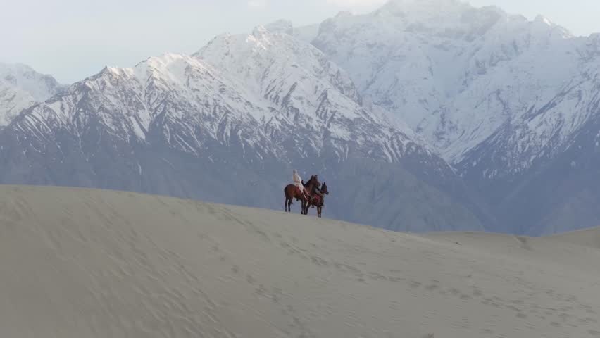 Horseback riders traverse the serene sand dunes against the backdrop of majestic snow-capped mountains in Pakistan
