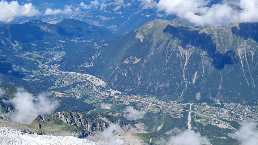 A video captured from a high altitude, showcasing a picturesque Alpine valley cradling a dense town (such as Chamonix or a similar resort)