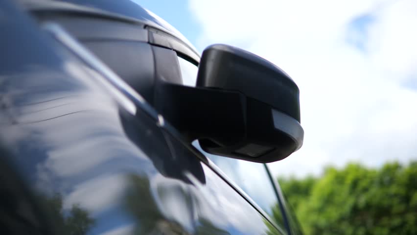 A car's side mirror elegantly unfolds in a smooth, automated motion, set against a soft-focus backdrop of a clear sky and rustling trees. 