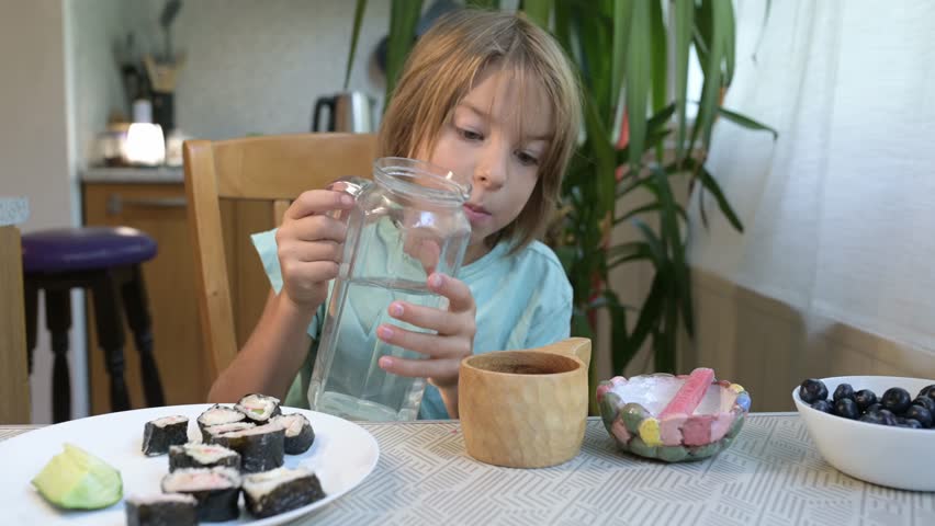 Young boy pouring water from a glass jug into a wooden cup while sitting at the kitchen table with sushi and blueberries
