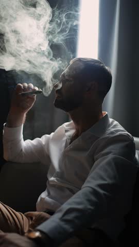 Elegant man in white shirt smoking cigar indoors, dramatic portrait of confident businessman exhaling smoke in low light, luxury lifestyle and relaxation concept