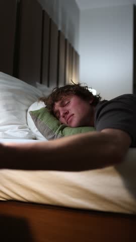 Vertical screen young man lying on bed late at night eating snacks from plate unhealthy habit of midnight binge casual atmosphere of home rest relaxation and everyday lifestyle indoors