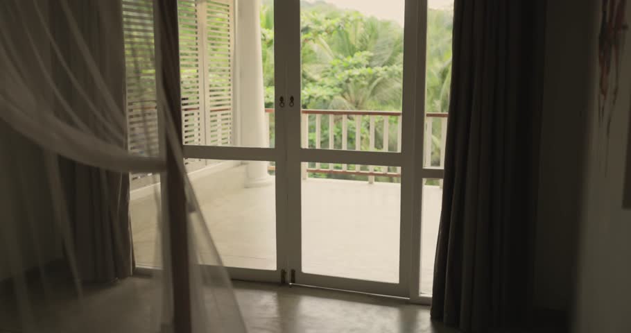 Stylish young woman in white summer dress and hat arrives at tropical resort room with suitcase, walking to balcony and enjoying lush jungle view. Perfect high quality travel and tourism promo.