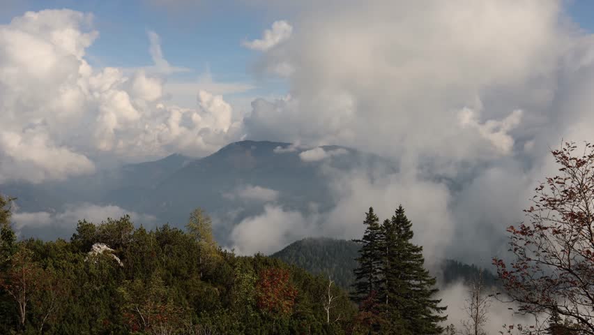 Majestic mountain landscape with towering clouds over misty alpine peaks and dark green forest trees blue sky opening distant valleys creating atmospheric wilderness scenery and travel mood