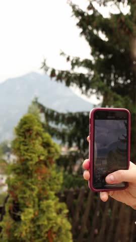 Vertical screen hand holds smartphone capturing scenic mountain landscape with tall green trees and misty peaks in background blending technology with nature in peaceful outdoor moment