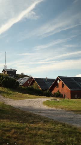 Vertical screen group of wooden mountain houses with solar panels on rooftops stands along winding gravel path under blue sky representing renewable energy use and eco friendly alpine living
