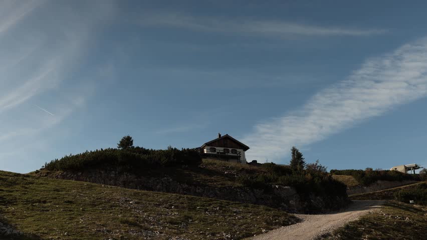 Mountain cabin with wooden roof stands on hilltop above rocky slope under clear blue sky with scattered clouds and winding dirt road leading uphill in alpine landscape