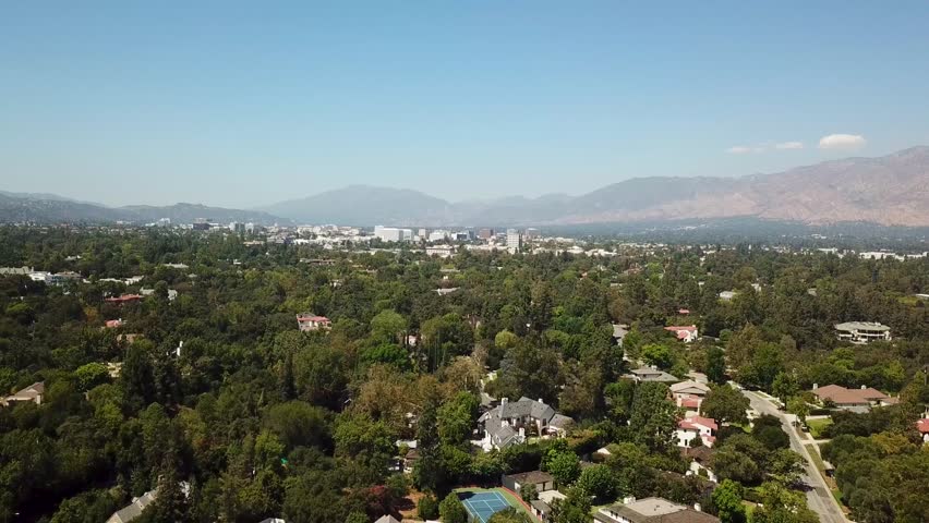 Smooth aerial pullback from above a leafy Pasadena neighborhood, widening to reveal a carpet of homes, tall trees, and the distant San Gabriel Valley skyline on a clear sunny day.