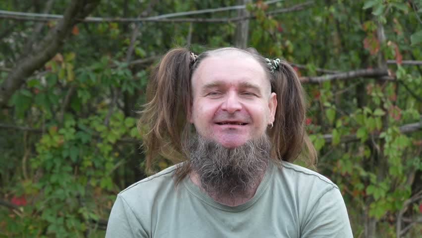 A bearded man with women's ponytails makes silly facial expressions and laughs in a humorous way during a staged outdoor shoot. Video captured in time-lapse at 50 fps.