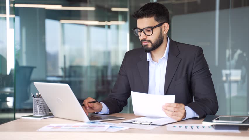 Busy businessman in formal suit is doing paperwork using computer while sitting at workplace in business office. Thoughtful financier working with documents, making a report, engaged in accounting