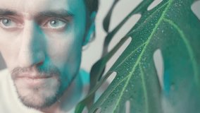 A young handsome man with beautiful blue eyes smiles playfully next to a large monstera leaf in soft teal lighting - Powered by Shutterstock - Get 15% off with code: PIKWIZARD15
