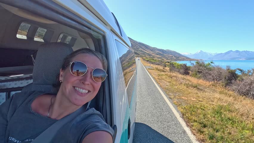 A girl sits in the passenger seat of a camper van admiring Lake Pukaki, South Island, New Zealand. The turquoise lake and mountains create a stunning backdrop on this scenic road trip.