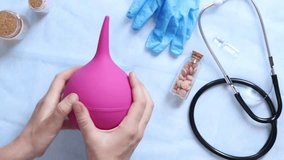 Doctor's hands holding a large pink enema on a table background with various objects, top view. Female doctor squeezing a pink rubber enema. Nurse or doctor's desk with a stethoscope, pills and gloves - Powered by Shutterstock - Get 15% off with code: PIKWIZARD15