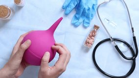 Doctor's hands holding a large pink enema on a table background with various objects, top view. Female doctor squeezing a pink rubber enema. Nurse or doctor's desk with a stethoscope, pills and gloves - Powered by Shutterstock - Get 15% off with code: PIKWIZARD15