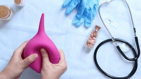 Doctor's hands holding a large pink enema on a table background with various objects, top view. Female doctor squeezing a pink rubber enema. Nurse or doctor's desk with a stethoscope, pills and gloves - Powered by Shutterstock - Get 15% off with code: PIKWIZARD15