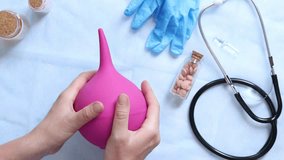 Doctor's hands holding a large pink enema on a table background with various objects, top view. Female doctor squeezing a pink rubber enema. Nurse or doctor's desk with a stethoscope, pills and gloves - Powered by Shutterstock - Get 15% off with code: PIKWIZARD15