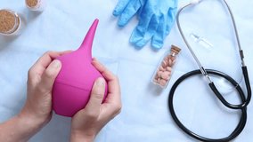 Doctor's hands holding a large pink enema on a table background with various objects, top view. Female doctor squeezing a pink rubber enema. Nurse or doctor's desk with a stethoscope, pills and gloves - Powered by Shutterstock - Get 15% off with code: PIKWIZARD15