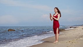 Young woman in red dress stands barefoot on sandy Baltic Sea shore, capturing selfie with clear blue sky and waves in background. Peaceful coastal setting highlights calm waters and natural beauty - Powered by Shutterstock - Get 15% off with code: PIKWIZARD15