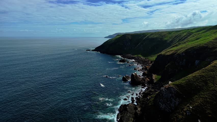 Waves washing along steep green cliffs on North Sea coast in Scotland. Swells gliding past rugged shoreline under partly cloudy sky. Foam trailing through rock formations beneath rolling hills