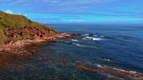 Waves lapping against red sandstone cliffs on North Sea coast in Scotland. Swells reaching weathered shoreline under clear blue sky. Tides washing over layered rocks near rugged headland. Currents - Powered by Shutterstock - Get 15% off with code: PIKWIZARD15