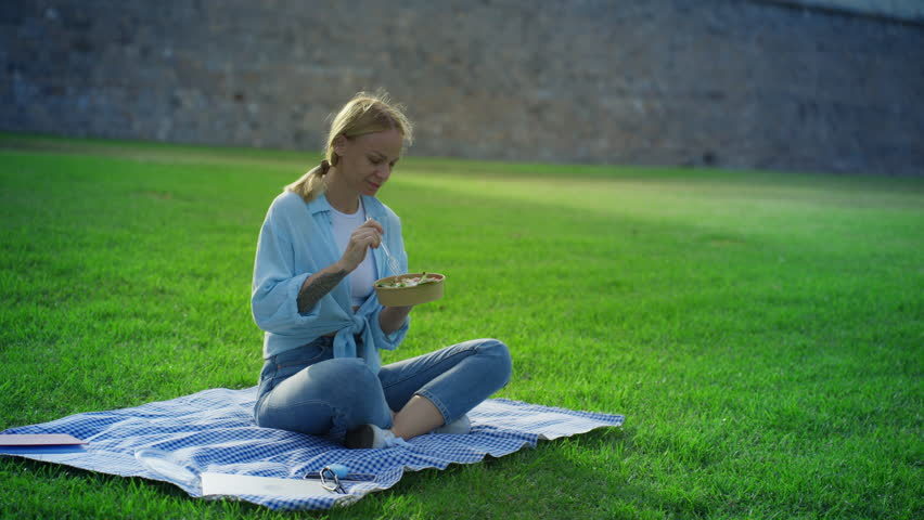 Young beautiful woman eating fresh green salad outdoors, sitting on plaid blanket on grass for healthy picnic lunch break, enjoy peaceful moment in sunlit city park. Concept picnic.Vegeterian