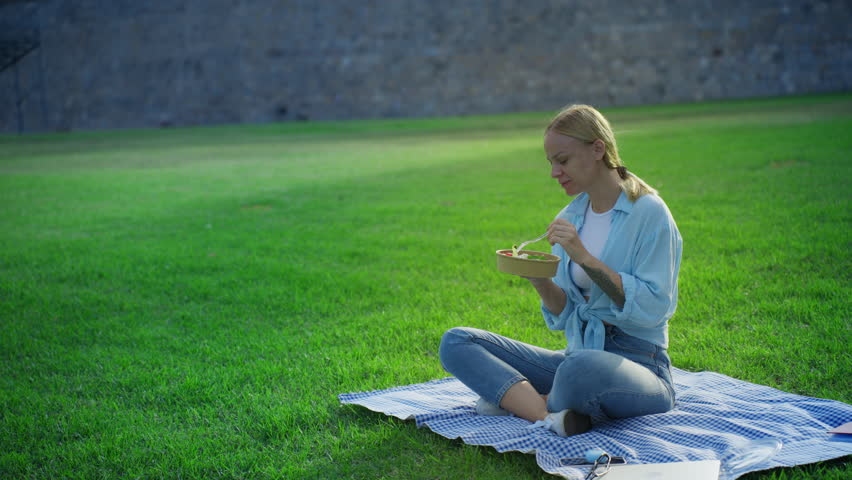 Youthful blonde woman sits on blue checkered blanket in sunny city park, smiling as she enjoy fresh vegetable salad during relaxed outdoor lunch on green grass. Concept picnic.Vegeterian