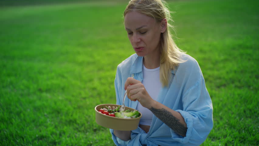 Young woman sits on lush green lawn in sunny city park, enjoy fresh salad from takeaway bowl, relaxed and smiling during healthy lunch outdoors. Concept picnic.Vegeterian