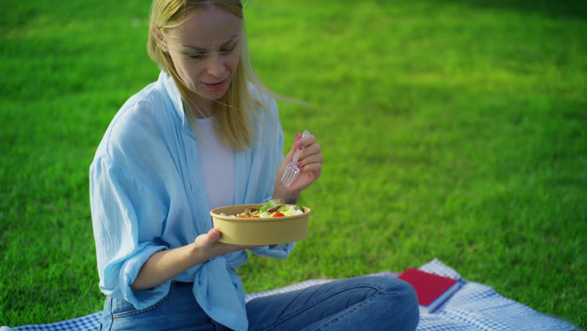 Pretty young woman sitting on checkered blanket on green grass enjoy fresh salad from takeaway bowl, have healthy vegetarian lunch during relaxing picnic in park.Concept picnic.Vegeterian