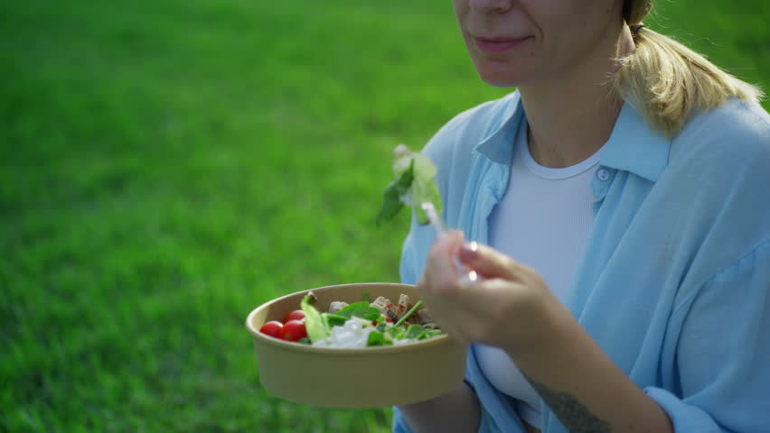 Close-up of smiling woman enjoy fresh vegetable salad with tomatoes and chicken from craft bowl, eating with fork while sitting on green lawn in park during her lunch break.Concept picnic.