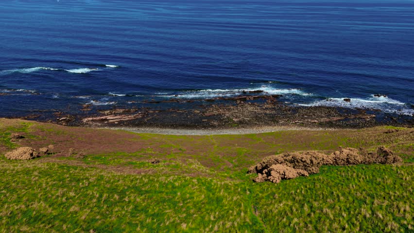 Waves rolling onto rocky shoreline along deep blue ocean. Sea meeting scattered stones at edge of grassy cliff. Surf crashing gently on coastal rocks near green slope. Ocean stretching calmly beyond