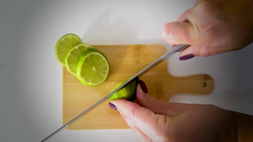 Close-up of female hand using a sharp knife to slice a fresh green lime into thin round pieces on a small wooden chopping board. Healthy citrus fruit preparation, perfect for cooking, drinks