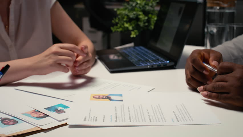 Close up of CV document evaluation in a corporate job interview, businesswoman asking african american candidate questions based on his past work experience and studies shown on paper. Camera A.
