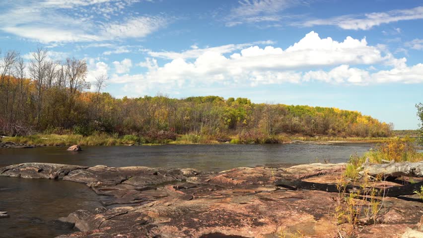 Calm river with rocky shore and autumn forest. Colorful trees with yellow and orange leaves under blue sky with clouds.