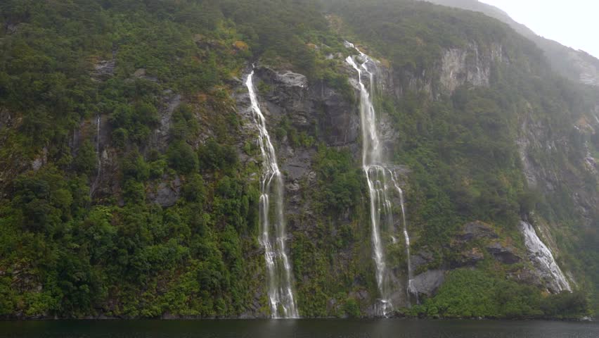 Sailing past twin waterfalls in Doubtful Sound, Fiordland National Park, New Zealand. Mist rises from the cascading water as the boat glides through the dramatic fjord landscape.
