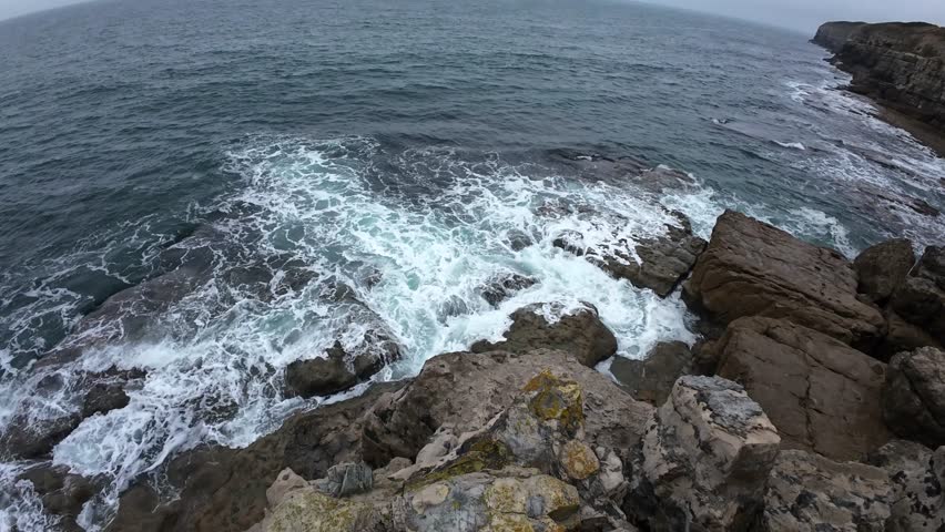 Dynamic aerial footage showcasing the rugged beauty of Dancing Ledge, Isle of Purbeck, Dorset. Waves crash against weathered rocks under overcast skies, moodily capturing natural coastal drama.