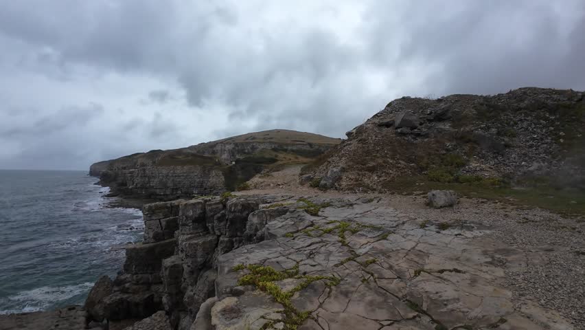 Dramatic aerial footage captures rugged rock formations and crashing waves on a moody, overcast day at the Isle of Purbeck, Dorset. The wide ocean view creates a serene yet powerful atmosphere.