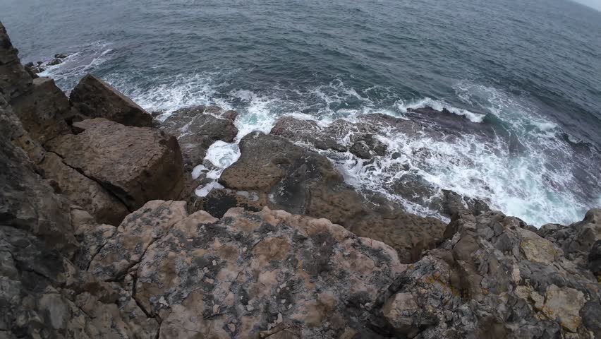 Drone video captures stunning aerial view of waves crashing on rustic rocks at Dancing Ledge, Isle of Purbeck. Dramatic lighting and rugged formations create a dynamic coastal scene.