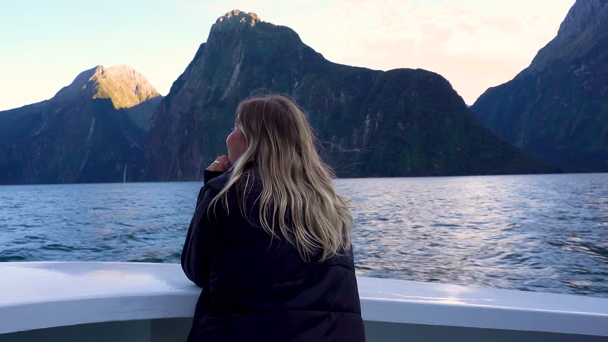 A woman stands at the front of a boat sailing through Milford Sound in Fiordland National Park, New Zealand. She is taking in the towering cliffs and waterfalls all around the fjord.