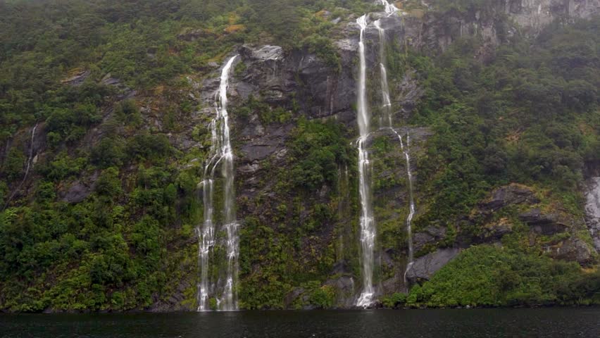 A boat glides through Doubtful Sound in Fiordland National Park, New Zealand, passing twin waterfalls. Mist rises from the cascading water as mountains tower along the fjord.