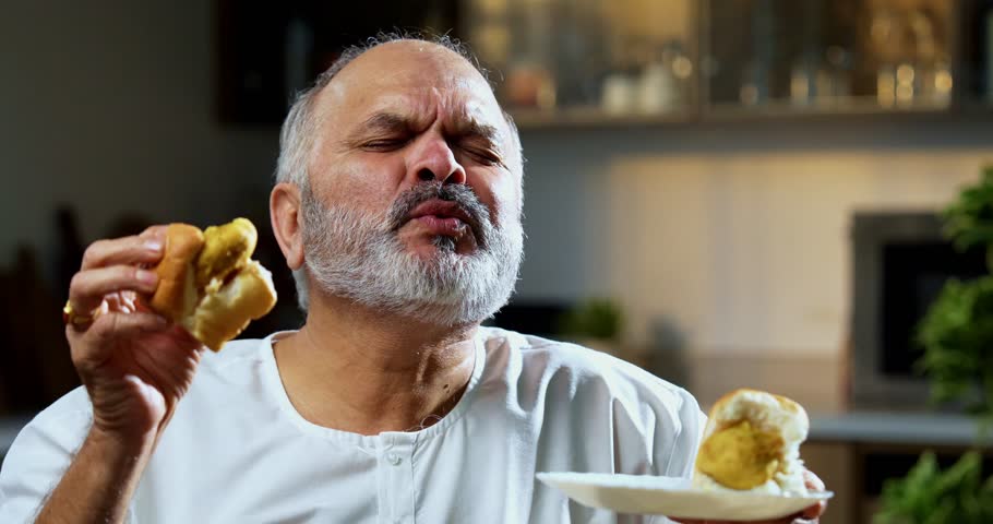 Senior Indian man eating vada pav with tea at home table, Asian foodie old man expressing immense joy and satisfaction while enjoying spicy Mumbai street food snack in modern Indian kitchen setup