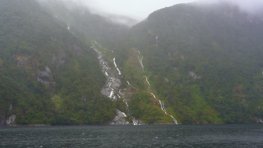 Waterfalls cascade from mountains on a misty, cloudy day in Doubtful Sound, Fiordland National Park, New Zealand. Mist rises as water flows into the fjord below.