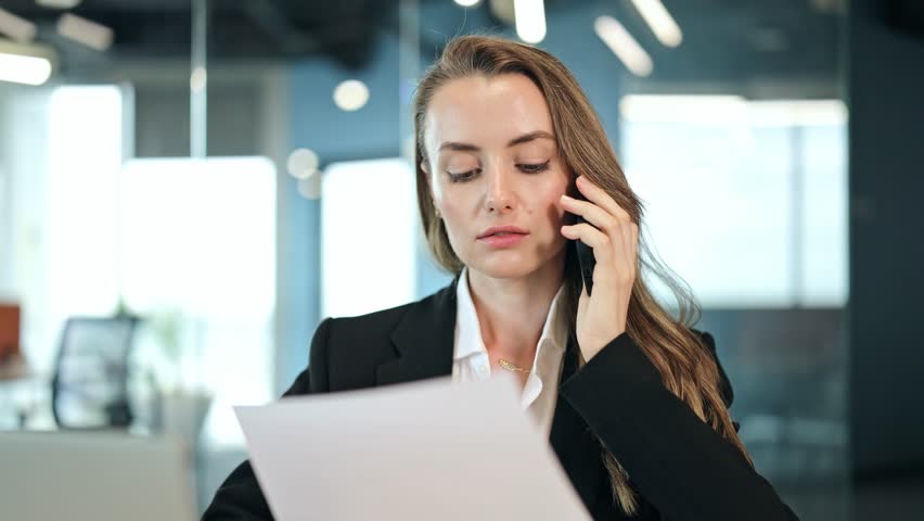 Focused businesswoman, dressed in black blazer and white shirt, in modern office. Serious businesswoman engages in phone call reviewing important documents, dedication and efficient communication.