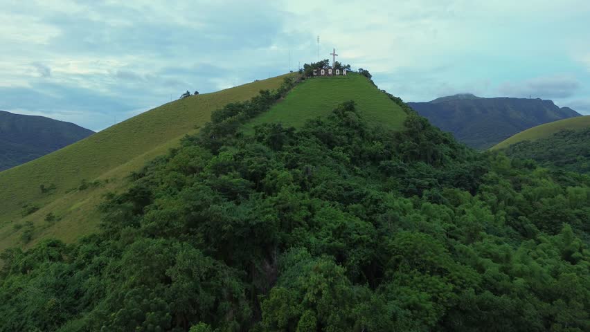 Drone rises toward Mount Tapyas and the hilltop cross above Coron town, Palawan. Grassy green slopes and the CORON sign lead up to the ridge under soft evening light and textured clouds.