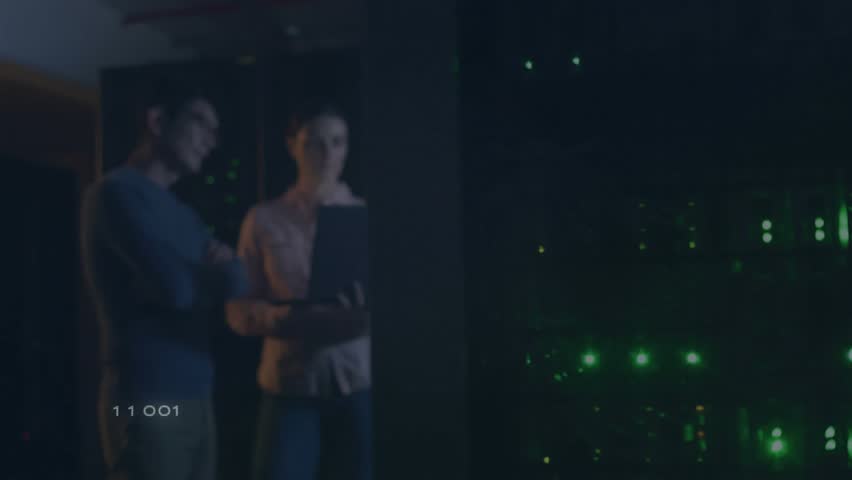 Female IT pro offering laptop to male engineer in data center showing binary overlays for analysis. Technology, cybersecurity, innovation, networking, digital, professional, collaborative - Powered by Shutterstock - Get 15% off with code: PIKWIZARD15