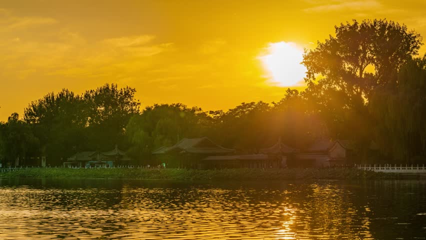 Golden sunset over Shichahai lake in Beijing, China with traditional architecture silhouetted against vibrant orange sky and water reflections during evening dusk.