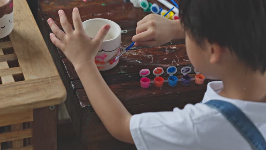 Asian Child painting ceramic cup with colorful paint on wooden table creative activity art supplies brush focused kid enjoying indoor hobby artistic expression leisure time craft project
