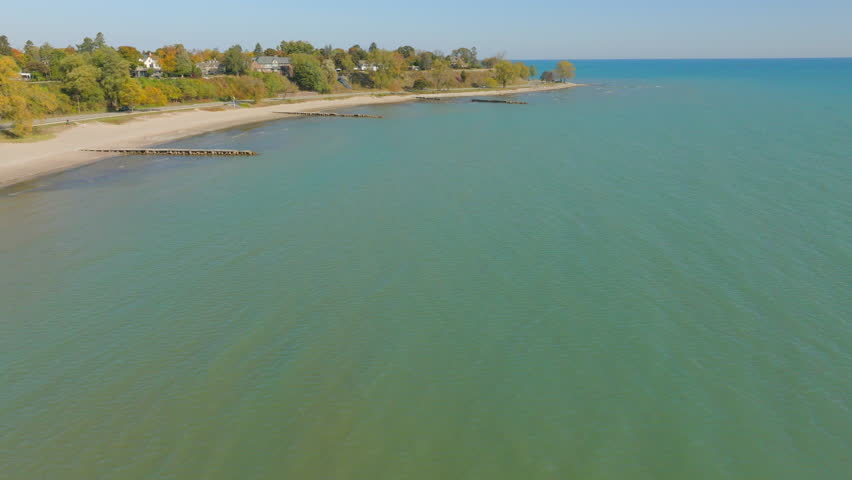 Drone aerial over bright blue water of Lake Michigan on a pretty autumn day toward the Sheboygan, Wisconsin shoreline with colorful trees and charming neighborhood houses.