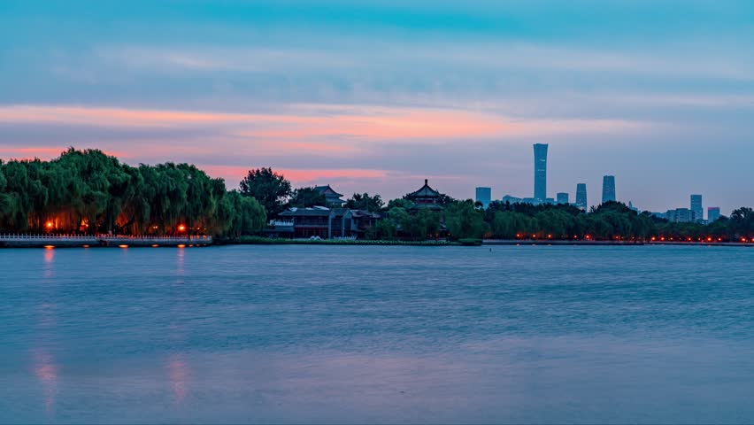 Beautiful morning timelapse view of Shichahai lake in Beijing, China with traditional architecture and modern skyline reflected in calm waters under colorful dawn sky.
