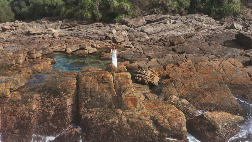 A cinematic aerial flyover of a woman in a flowing white dress standing on a rugged cliff as ocean waves crash below. Ideal for nature, travel, and dramatic landscape concepts. 